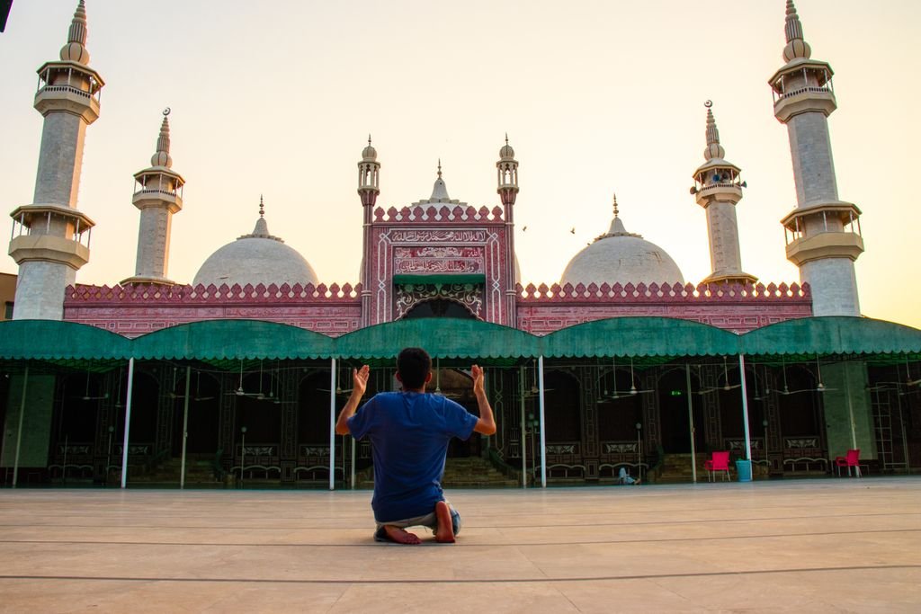 A man kneels in prayer outside a mosque in Faisalabad during sunset, showcasing Islamic architecture.